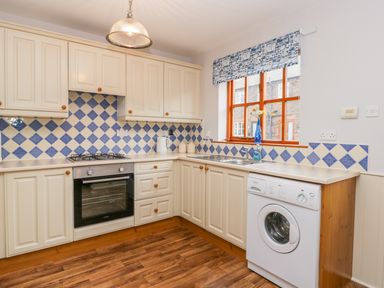 A kitchen with white cabinets blue and white tiled backsplash a gas stove oven washing machine and a window with a blue patterned blind at Little Venice in Whitby