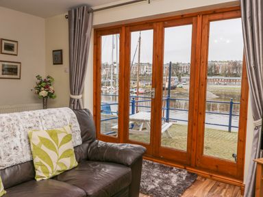 A living room with a leather sofa flowers on a stand and large wooden framed glass doors showing a harbor at Little Venice in Whitby