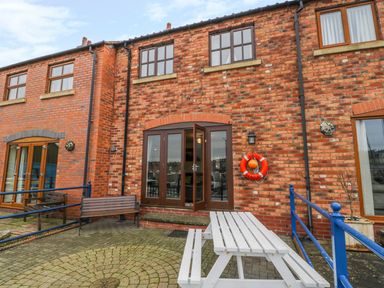 An outdoor paved patio area with a white picnic table bench and wooden bench seating attached to a red brick building with brown framed windows and a life buoy on the wall at Little Venice in Whitby