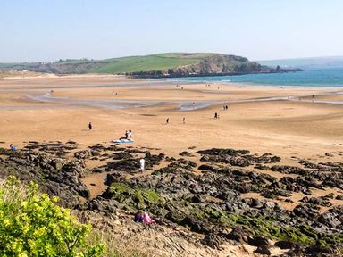 A beach scene with sand, rocks, scattered people, green hills, and the sea at Quarterdeck in Bigbury-on-Sea near Modbury