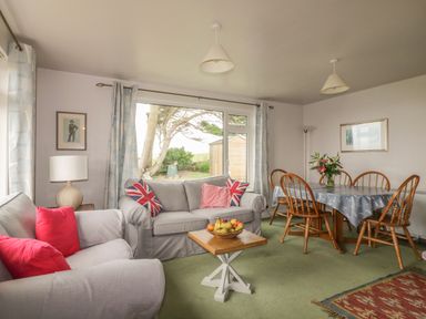 A living room with sofas, a wooden dining table with chairs, a lamp, and a window with curtains at Quarterdeck in Bigbury-on-Sea near Modbury