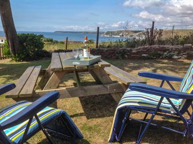 An outdoor picnic table with a bottle and two glasses on a tray and two striped lounge chairs with armrests at Quarterdeck in Bigbury-on-Sea near Modbury