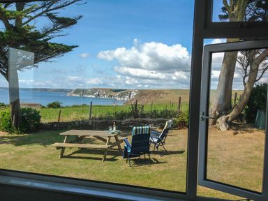 A garden with a wooden picnic table and two lawn chairs overlooking cliffs and the sea at Quarterdeck in Bigbury-on-Sea near Modbury