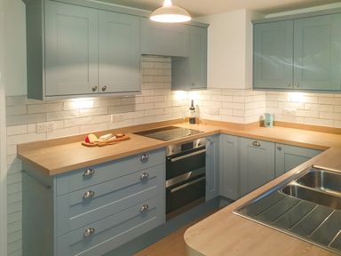 A kitchen with blue cabinets wooden countertops an electric stove a double oven a stainless steel sink and a wooden board with cheese and grapes at Quarterdeck in Bigbury-on-Sea near Modbury