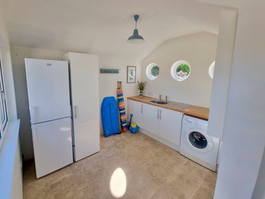 A utility room with a refrigerator, cabinets, sink, washing machine, and surfboards at Quarterdeck in Bigbury-on-Sea near Modbury