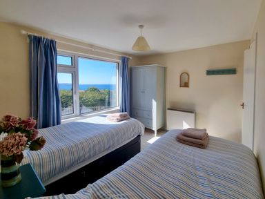A bedroom with two single beds blue striped bedding a wardrobe and a window with a sea view at Quarterdeck in Bigbury-on-Sea near Modbury