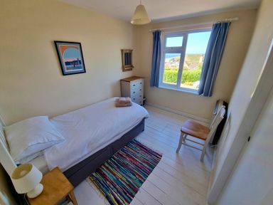 A bedroom with a single bed a wooden chair a chest of drawers a window with blue curtains and a colorful rug on the floor at Quarterdeck in Bigbury-on-Sea near Modbury
