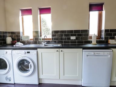 A kitchen area with a washing machine, dryer, dishwasher, sink, countertop appliances, and three windows with purple blinds at Seascape in Bridlington