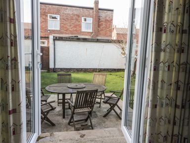 A backyard with a round wooden table and six wooden chairs viewed from open double glass doors with curtains at Seascape in Bridlington