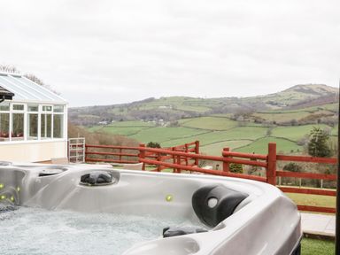 An outdoor hot tub with bubbles next to a conservatory and a wooden fence overlooking green hills at Bryn Derw Holiday Cottage in Eglwysbach near Llanrwst and Conwy