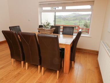 A dining area with a wooden table and eight brown chairs near a window with a view of green hills at Bryn Derw Holiday Cottage in Eglwysbach near Llanrwst and Conwy