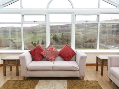 A seating area with a sofa and cushions in front of large windows showing a rural landscape at Bryn Derw Holiday Cottage in Eglwysbach near Llanrwst and Conwy