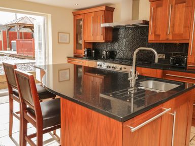 A kitchen with wooden cabinets a black countertop two chairs and an open door leading outside at Bryn Derw Holiday Cottage in Eglwysbach near Llanrwst and Conwy