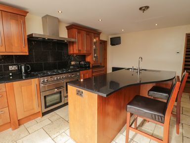 A kitchen with wooden cabinets a black countertop gas stove oven and two bar stools at Bryn Derw Holiday Cottage in Eglwysbach near Llanrwst and Conwy