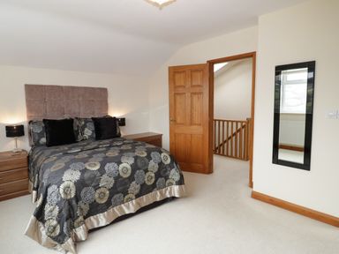 A bedroom with a bed with patterned bedding two bedside tables lamps an open wooden door a staircase and a wall mirror at Bryn Derw Holiday Cottage in Eglwysbach near Llanrwst and Conwy
