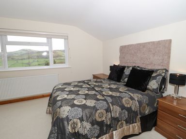 A bedroom with a double bed with patterned bedspread and bedside tables with a lamp and window showing a view of hills at Bryn Derw Holiday Cottage in Eglwysbach near Llanrwst and Conwy