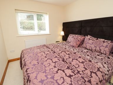 A bedroom with a large bed covered in a floral patterned bedspread and pillows next to a window with blinds and a bedside table with a lamp at Bryn Derw Holiday Cottage in Eglwysbach near Llanrwst and Conwy
