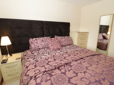 A bedroom with a bed with floral bedding a bedside table with a lamp and remote controls a chest of drawers and a full length mirror at Bryn Derw Holiday Cottage in Eglwysbach near Llanrwst and Conwy