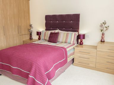 A bedroom with a bed with purple headboard and striped bedding two bedside tables with lamps and flowers at Bryn Derw Holiday Cottage in Eglwysbach near Llanrwst and Conwy