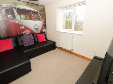 A living room with a black leather sofa with cushions a large wall mural of a red and white Volkswagen van a window with a radiator below and a television on a stand Bryn Derw Holiday Cottage in Eglwysbach near Llanrwst and Conwy