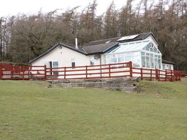 A single-story house with a glass conservatory and wooden fence on a grassy hill with trees in the background at Bryn Derw Holiday Cottage in Eglwysbach near Llanrwst and Conwy