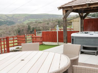 An outdoor patio area with a round table, wicker chairs, a hot tub under a wooden gazebo, and a fenced grassy yard with hills in the background at Bryn Derw Holiday Cottage in Eglwysbach near Llanrwst and Conwy