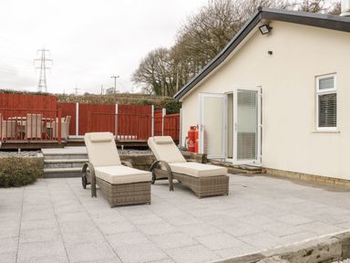 An outdoor patio area with two cushioned lounge chairs and an open door leading into a cream-colored building with a red wooden fence and outdoor dining table in the background at Bryn Derw Holiday Cottage in Eglwysbach near Llanrwst and Conwy