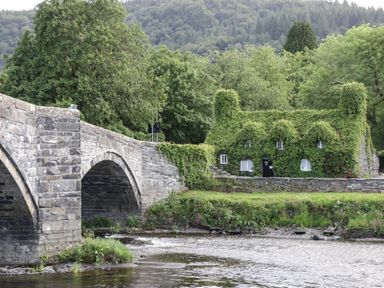 Stone bridge over a river with a house covered in ivy and trees in the background at Bryn Derw Holiday Cottage in Eglwysbach near Llanrwst and Conwy