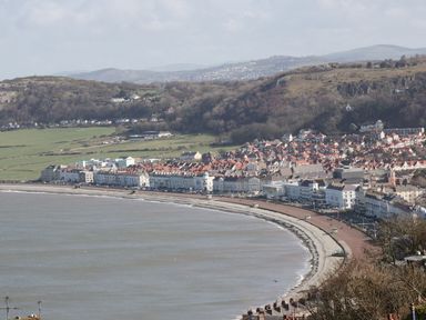 A coastal town with a curved seafront promenade backed by rows of buildings and hills in the background at Bryn Derw Holiday Cottage in Eglwysbach near Llanrwst and Conwy