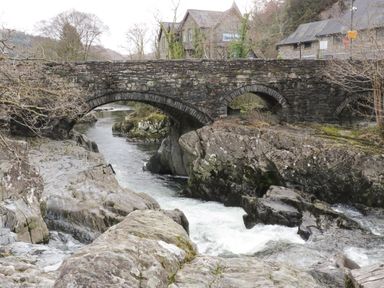 A stone arch bridge over a river with rocks and buildings in the background at Bryn Derw Holiday Cottage in Eglwysbach near Llanrwst and Conwy