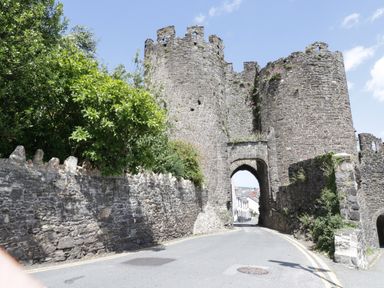 A stone castle gate with two round towers on a road with stone walls and greenery under a blue sky