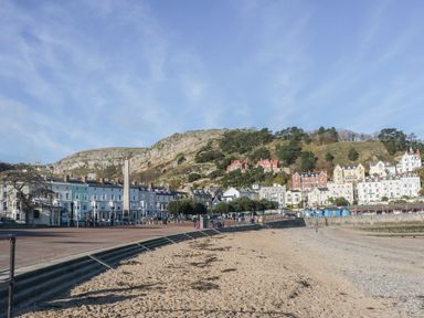 A sandy beach with a promenade and a row of buildings at the base of a rocky hill
