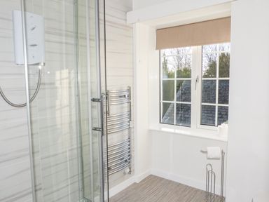 A bathroom with a glass enclosed shower, chrome towel radiator, window with blinds, and toilet paper holder at The Nook in Bowness-On-Windermere