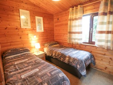 A bedroom with two single beds a nightstand with a lamp two framed pictures on wood panel walls and a window with striped curtains at Hawthorne Lodge in Liverton near Danby