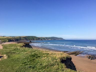 A coastal cliff with grassy top overlooking a sandy beach and ocean at Atlantic Reach in Widemouth Bay