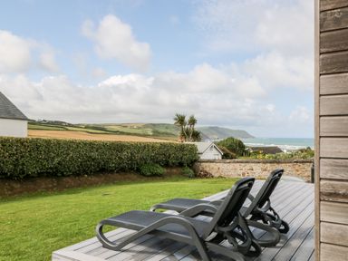 Two lounge chairs on a wooden deck overlooking a lawn with a hedge and coastal landscape at Atlantic Reach Widemouth Bay
