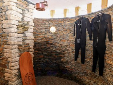 A stone room with a wooden surfboard and two wetsuits hanging on the wall at Atlantic Reach in Widemouth Bay