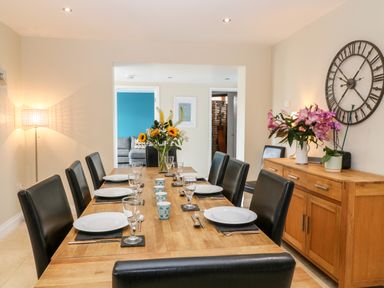 A dining room with a wooden table set with plates glasses and cutlery black chairs a sideboard with flowers and a large wall clock at Atlantic Reach in Widemouth Bay
