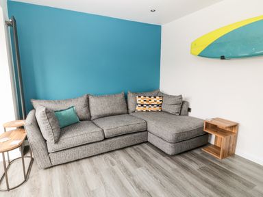 A living room with a grey corner sofa two wooden side tables and a surfboard on the wall at Atlantic Reach in Widemouth Bay