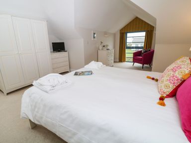 A bedroom with a large bed pillows a wardrobe a dresser TV and a pink chair by the window at Atlantic Reach in Widemouth Bay
