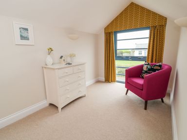 A room with a white chest of drawers and a pink armchair next to a window with yellow curtains at Atlantic Reach in Widemouth Bay