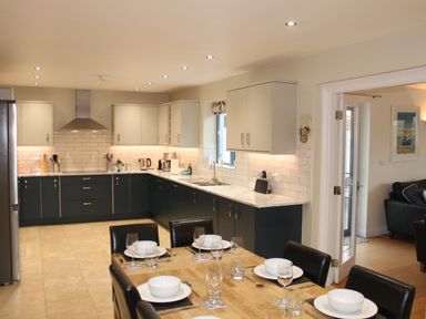 A kitchen with dark lower cabinets and light upper cabinets a dining table set with plates bowls and glasses at Atlantic Reach in Widemouth Bay
