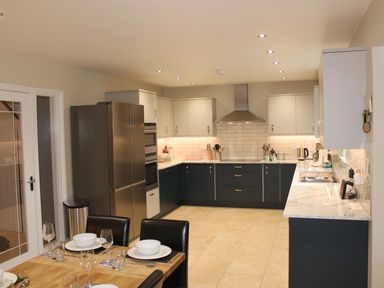 A kitchen with dark lower cabinets and light upper cabinets a stainless steel refrigerator and a dining table with set plates and glasses at Atlantic Reach in Widemouth Bay