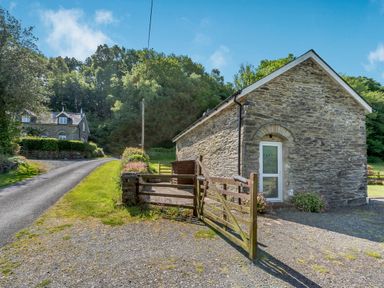 A stone building with a wooden gate and gravel driveway near a road and trees at Cwmdu Cottage in Cwm-Cou near Newcastle Emlyn