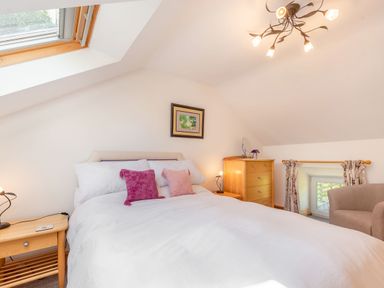 A bedroom with a bed white bedding wooden nightstands chest of drawers and beige armchair at Cwmdu Cottage in Cwm-Cou near Newcastle Emlyn
