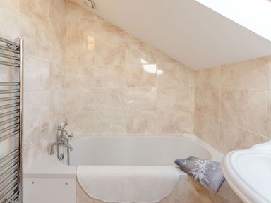 A bathroom with a bathtub under a slanted ceiling with beige tiled walls at Cwmdu Cottage in Cwm-Cou near Newcastle Emlyn