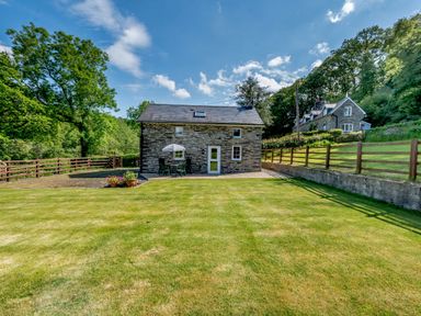 A stone cottage with lawn and outdoor table with umbrella surrounded by trees and fencing at Cwmdu Cottage in Cwm-Cou near Newcastle Emlyn