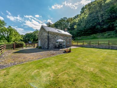 A stone cottage with a patio table and umbrella in a fenced yard with grass and trees at Cwmdu Cottage in Cwm-Cou near Newcastle Emlyn