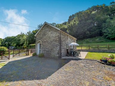 A stone cottage with a patio table and chairs under an umbrella in a grassy garden at Cwmdu Cottage in Cwm-Cou near Newcastle Emlyn
