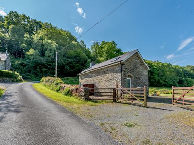 A stone cottage with wooden gates next to a road and trees in the background at Cwmdu Cottage in Cwm-Cou near Newcastle Emlyn